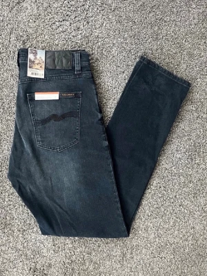 Mörkblå jeans från Nudie Jeans - Snygga mörkblå jeans från Nudie Jeans med klassisk femficksdesign och diskret slitning framtill. Jeansen har smal passform och är tillverkade i mjuk denim med stretch. Bakfickan har Nudie Jeans ikoniska broderi. Storleken är 31/34, men sitter lite mindre som 30/32