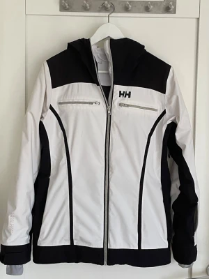Svartvit jacka från Helly Hansen, skidbyxor från 8848 Altitude - Snygg svartvit skidjacka från Helly Hansen i strl M (38) med huva. Två bröstfickor med dragkedja framtill och HH-logga på bröstet och ryggen. Jackan är vind-och vattentät.                                        Snygga vita skidbyxor (dam), strl M