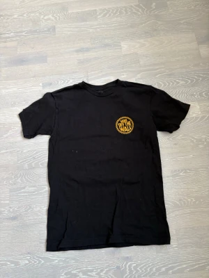 Svart Vans Off The Wall t-shirt - Svart t-shirt från Vans med gul 'Off The Wall'-logga på bröstet i ett runt märke med schackrutigt mönster runt om. Klassisk passform och rund halsringning. Perfekt för dig som gillar streetwear och enkel stil.