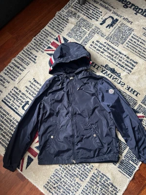 Mörkblå Moncler vindjacka med huva - Snygg mörkblå vindjacka från Moncler med huva och dragkedja. Jackan har vita och röda ränder på huvan samt Moncler-logga på ärmen. Tillverkad i lätt och glansigt material som står emot vind. Perfekt för dig som vill ha en stilren och sportig look. S/M