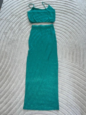 Glittrigt turkost set med kjol och topp - Säljer ett glittrigt turkost set med en croppad topp med tunna axelband och en lång, figurnära kjol. Materialet är stretchigt och skimrande, perfekt för dig som vill sticka ut. Märke saknas på plagget.