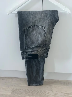 Grå raka jeans med stor bakficka - Säljer ett par grå jeans med rak passform och en stor bakficka. Jeansen har en klassisk femficksdesign och är tillverkade i denim. Perfekta för en avslappnad och trendig look.