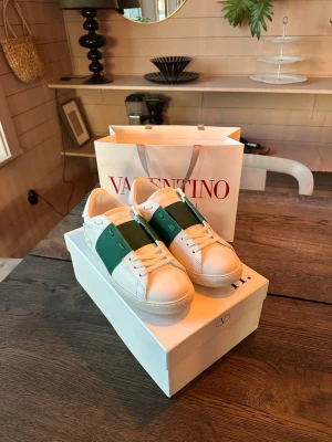 Valentino vita sneakers med grön detalj - Snygga vita sneakers från Valentino med en bred grön remsa över sidan och klassiska nitar på hälen. Skorna har lågt skaft, vita snören och är tillverkade i slätt läder. Perfekta för dig som vill ha en stilren men ändå unik look.
