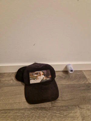 Svart truckerkeps med Marilyn Monroe - Säljer en svart truckerkeps med mesh på sidorna och ett stort tygmärke framtill med ett ikoniskt foto av Marilyn Monroe. Kepsen har böjd skärm och justerbar snapback baktill. Perfekt för dig som gillar retrostil och popkultur.