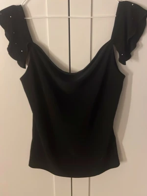 Svart topp med volangärmar och pärlor - Snygg svart topp med volangärmar dekorerade med små pärlor. Toppen har en mjuk, draperad urringning och är tillverkad i ett lätt, följsamt material. Perfekt för dig som vill ha något extra till outfiten.