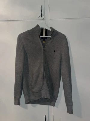 Grå ziptröja från Polo Ralph Lauren - Snygg grå ziptröja från Polo Ralph Lauren med hög krage och ribbade muddar. Tröjan har dragkedja framtill
