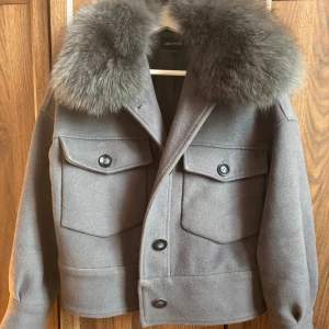 Meotine jacka - Snygg grå jacka med stor fluffig krage i matchande grått. Jackan har två stora bröstfickor med knappar och en rak, boxig passform. Perfekt för dig som vill ha en statementjacka med lyxig känsla och cool look.