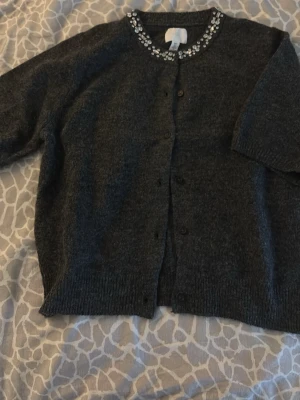 Mörkgrå kofta med strass från H&M - Mysig mörkgrå kofta från H&M med rund halsringning och dekorativa strassdetaljer längs kragen. Koftan har knäppning framtill och ribbade muddar vid ärmslut och nederkant. Perfekt för dig som gillar lite extra bling i vardagen.