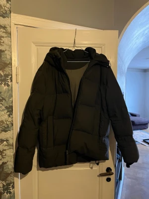 Svart pufferjacka från A Day's March - Snygg svart pufferjacka från A Day's March med huva och dragkedja. Jackan har stora quiltade partier och innerficka med dragkedja. Yttermaterial i polyester och fyllning av återvunnen polyester. Perfekt för kalla vinterdagar.