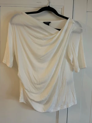 Vit draperad topp från Lindex - Snygg vit topp från Lindex med draperad front och korta ärmar. Tillverkad i mjukt, stretchigt material som ger en skön passform. Perfekt för dig som gillar en stilren men ändå unik look. Knappt använd!