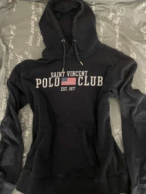Svart hoodie Saint Vincent Polo Club - Svart hoodie från Saint Vincent Polo Club med tryckt logga och amerikansk flagga på bröstet. Klassisk känguruficka framtill och justerbar huva med snören. Tillverkad i mjukt material, perfekt för en avslappnad och sportig stil.