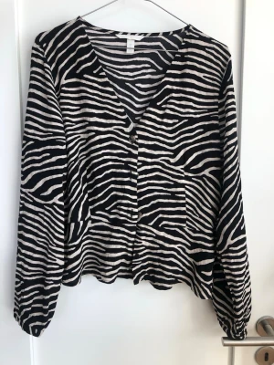 Zebramönstrad blus med knappar - Snygg blus med svartvitt zebramönster och v-ringning. Blusen har långa ärmar med resår vid ärmslut och knäppning framtill. Tillverkad i ett lätt och luftigt tyg som ger en avslappnad look. Liten i storleken och skulle säga mer som S