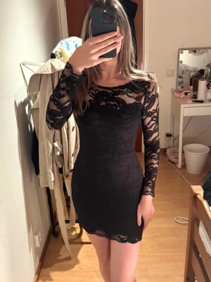 Svart spetsklänning med lång ärm - Säljer en svart, figurnära spetsklänning med långa ärmar och rundad hals. Klänningen har transparenta spetsdetaljer upptill och på ärmarna, samt en kort, kroppsnära passform. Har använt den en gång!