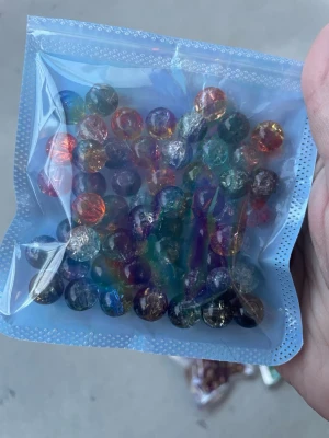 Flerfärgade glaspärlor i påse - Säljer en försluten påse med runda glaspärlor i olika färger som blå, grön, orange och lila. Pärlorna har en glansig yta och vissa har en spräcklig effekt inuti. Perfekt för pyssel, smyckestillverkning eller dekoration.