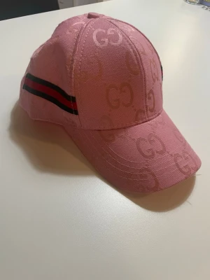 Rosa keps från Gucci med GG-mönster - Säljer en rosa keps från Gucci med det klassiska GG-mönstret över hela kepsen. Kepsen har en böjd skärm och en svart-röd rand på sidan som ger en sportig touch. Tillverkad i tygmaterial och perfekt för dig som vill sticka ut.