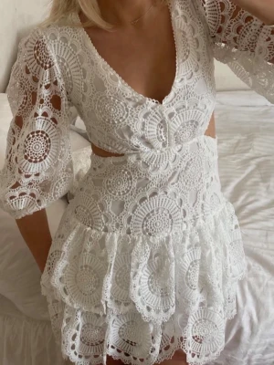 Vit spetsklänning med volanger - Superfin vit kortklänning i spets med v-ringning, trekvartsärm och volangdetaljer. Klänningen har öppen rygg och fina broderade mönster i hela tyget. Perfekt för sommarens alla tillfällen och ger en romantisk vibe.