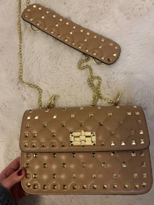 Beige axelremsväska med nitar - Snygg beige handväska i quiltat läder med guldfärgade nitar och kedja. Väskan har ett dekorativt spänne framtill och avtagbar axelrem med matchande nitar. Perfekt för dig som gillar edgy detaljer och trendig design. Aldrig använd. Hör av er vid frågor ❤️❤️