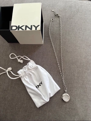 DKNY silverfärgat halsband med cirkelhänge  - Snyggt halsband från DKNY i silverfärg med ett runt hänge täckt av gnistrande stenar. Kedjan är tunn och elegant, och halsbandet kommer med original ask och vit tygpåse med DKNY-logga. Perfekt accessoar för att lyfta din outfit. Använt ett fåtal gånger 