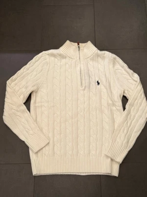 Vit kabelstickad tröja Polo Ralph Lauren - Kabelstickad vit tröja från Polo Ralph Lauren med halv dragkedja och hög krage. Tröjan har långa ärmar, ribbade muddar och den klassiska broderade loggan på bröstet. Perfekt för dig som gillar stilrena och tidlösa plagg.
