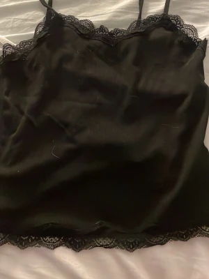 Svart spetskantad linnetopp - Snygg svart linnetopp med tunna axelband och dekorativ spets längs både urringning och nederkant. Materialet har en lätt glansig finish som ger en lyxig känsla. Perfekt att styla lager på lager eller som den är.