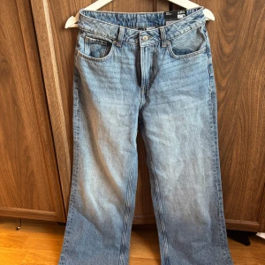 Blå raka jeans från H&M - Ett par klassiska blå jeans från H&M med rak passform och hög midja. Jeansen har fem fickor, knappgylf och är tillverkade i denim. Perfekta för en avslappnad och trendig look.Aldrig använda