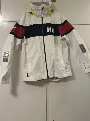 Vit vindjacka Helly Hansen - Snygg vit vindjacka från Helly Hansen med svart och röd rand över bröst och armar samt gul huva. Jackan har dragkedja framtill, två sidofickor med dragkedja och HH-logga på bröstet och huvan. Perfekt för blåsiga dagar och streetwear-stil. NYPRIS 3999 KR