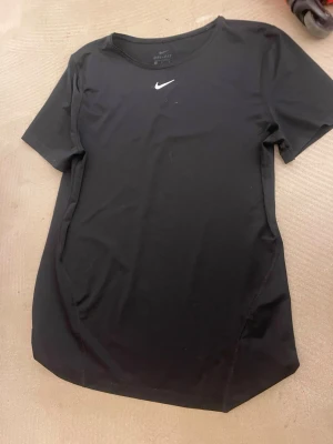 Svart Nike tränings t shirt  - Snygg svart t-shirt från Nike med klassisk vit logga på bröstet. Tillverkad i funktionsmaterialet Dri-Fit som andas och passar perfekt till träning. T-shirten har rund halsringning och korta ärmar för en sportig look.