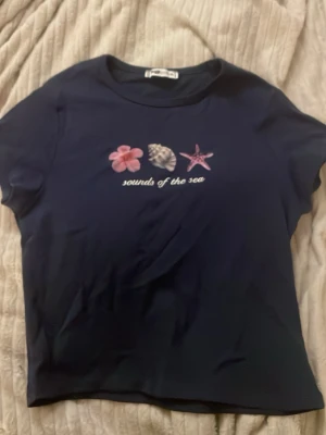 Mörkblå t-shirt med havsmotiv - Mörkblå t-shirt från FB Sister med tryck av en blomma, snäcka och sjöstjärna samt texten 'sounds of the sea' på bröstet. Klassisk rund halsringning och korta ärmar. Perfekt för dig som gillar strandvibbar.