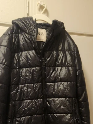 Svart pufferjacka från Zara - Snygg svart pufferjacka från Zara med huva och dragkedja framtill. Jackan har quiltad design och två sidofickor med dragkedja. Tillverkad i ett glansigt, lätt material som håller värmen under kalla dagar. Perfekt för dig som vill ha en stilren och praktisk vinterjacka.