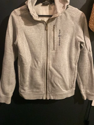 Ljusgrå hoodie från Sail Racing - Ljusgrå hoodie från Sail Racing med dragkedja framtill och en bröstficka med dragkedja. Märkets logga syns på bröstet. Hoodien har huva med dragsko och är tillverkad i mjukt, fleeceliknande material. Perfekt för chill dagar eller när du vill ha en clean look.