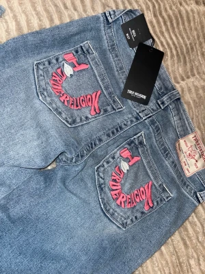 True Religion blå jeans med brodyr - Säljer ett par blå jeans från True Religion med ikonisk rosa brodyr på bakfickorna. Jeansen är bootcut och klassisk femficksdesign. Prislappar är kvar och de är endast testade 💕