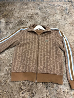 Brun mönstrad ziptröja från Lacoste - Snygg brun ziptröja från Lacoste med geometriskt mönster och klassisk krokodil-logga på bröstet. Tröjan har ribbade muddar, hög krage och blåvita ränder längs ärmarna. Perfekt för en sportig och trendig look.