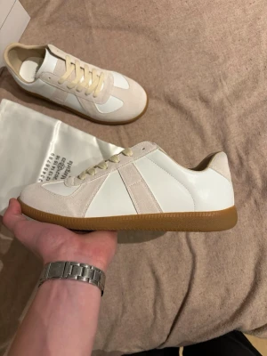 Maison Margiela Replica sneakers beige - Snygga Maison Margiela Replica sneakers i beige och vitt med klassisk gummisula. Ovandelen är i en mix av mocka och läder, med vita paneler och beige detaljer. Skorna har låga snitt och matchande beige skosnören. Perfekta för en clean och stilren look. Säljer de då de inte passade mig