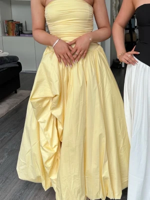 Gul axelbandslös balklänning - Snygg axelbandslös långklänning i ljusgul färg med voluminös kjol och draperad överdel. Klänningen har en ballongliknande form och slits och är perfekt för bal eller festliga tillfällen. 