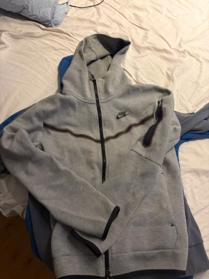Grå hoodie från Nike med dragkedja - Snygg grå hoodie från Nike med hel dragkedja och svart logga på bröstet. Hoodien har en huva, svarta detaljer vid ärmslut och fickor samt en dekorativ linje över bröstet. Perfekt för en sportig och avslappnad stil.