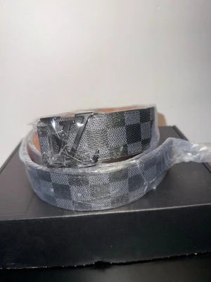 Svart Lv Bälte - Snyggt bälte från LV med klassiskt svart och grått rutmönster. Bältet har en stor LV-logga i metall på spännet och insidan är i ljusbrunt skinn. Perfekt accessoar för att lyfta din outfit.110CM verktyg för att justera storlek följer med 