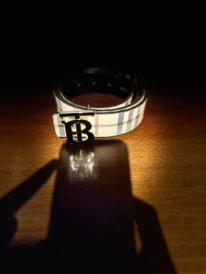 Snyggt bälte - Säljer ett snyggt rutig bälte  med klassiskt logospänne i metall. Bältet har beige, svart, röd och blå färgtoner och är tillverkat i skinn med typiskt mönster. Perfekt för att lyfta din outfit med en ikonisk accessoar.