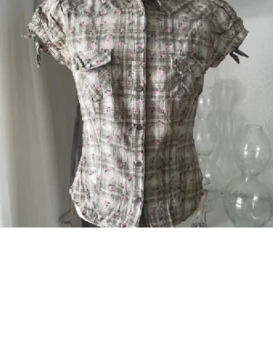 Rutig kortärmad skjorta med blommor - Kortärmad skjorta med rutigt mönster i beige och grått, dekorerad med små rosa blommor. Skjortan har knappar framtill, bröstfickor med lock och snörning på ärmarna för extra detalj. Perfekt för dig som gillar en söt och avslappnad stil.