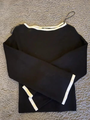Svart långärmad topp med vit kant - Svart långärmad topp från gina med vit kant längs halsringning och ärmslut. Enkel och stilren design med rak passform, perfekt att matcha med jeans eller kjol.