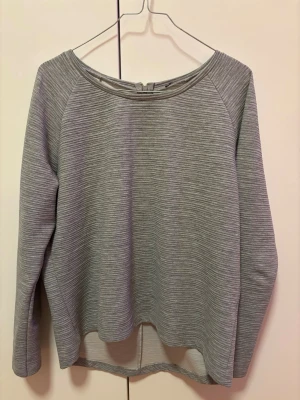 Grå långärmad tröja med struktur stl S-M - En stilren grå tröja med diskret strukturmönster och rundad halsringning. Tröjan har längre bakstycke och långa ärmar. Perfekt till jeans eller kjol för en enkel men snygg look. Storlekslapp bortklippt, passar en S-M
