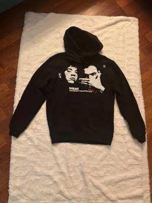 Svart hoodie M&M Tour Part 2 - Svart hoodie med stor huva och tryck fram av ett ansikte samt texten 'M&M WE ARE NOT THE SAME PART 2'. På ryggen finns turnélistor med städer och datum i vitt och orange. Hoodien har ribbade muddar och är i mjukt bomullsmaterial. Perfekt för dig som gillar streetwear och unika plagg. Inköpt på gröna Lund i juni 2025 för 650 sek.