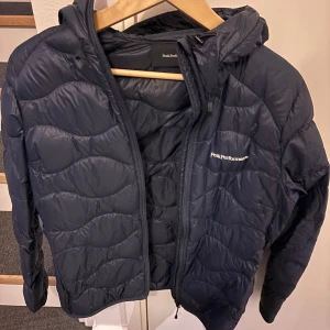 Marinblå lättviktsdunjacka Peak Performance - Snygg marinblå lättvikts dunjacka från Peak Performance med huva och dragkedja framtill. Jackan har diskret logga på bröstet och quiltad design som ger en modern look. Perfekt för kalla dagar och enkel att matcha med olika outfits.