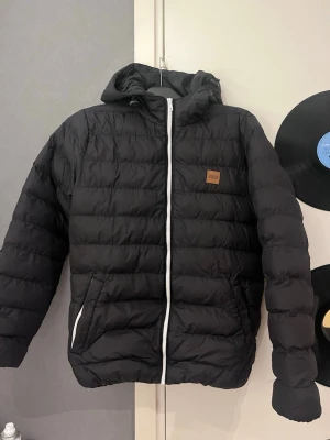 Svart dunjacka med huva från Urban - Säljer en svart dunjacka från Urban med huva och vita dragkedjor. Jackan har två fickor framtill och quiltad design. Perfekt för kalla dagar och har en stilren look med diskret logga på bröstet.