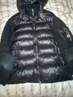 Svart dunjacka med huva från Moncler - Snygg svart dunjacka från Moncler med stickade ärmar och huva. Jackan har quiltad front, dragkedja och en ficka med Moncler-logga på ärmen. Perfekt för kalla vinterdagar och riktigt trendig look.