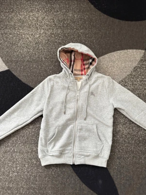 Burberry zip - Snygg grå zip hoodie från Burberry med klassiskt rutigt foder i huvan och broderad logga på bröstet. Hoodien har dragsko i huvan, två stora fickor fram och ribbade muddar. Perfekt för en chill och stilren look.