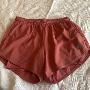Roströda träningsshorts - Säljer ett par roströda träningsshorts från Craft. De har elastisk midja och en sportig design med rundade kanter. Perfekta för löpning eller gymmet. Logga i silver på ena benet. Lätta och bekväma att bära.
