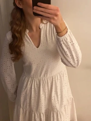 Vit långärmad klänning med spetsdetaljer - Söt vit klänning med v-ringning och lång ärm. Klänningen har broderade spetsdetaljer och lätt volang i kjolen. Perfekt för sommarens alla tillfällen och ger en romantisk känsla. Tillverkad i bomull för en luftig och bekväm känsla.