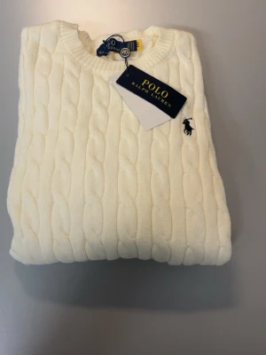Vit kabelstickad tröja Polo Ralph Lauren - Kabelstickad vit tröja från Polo Ralph Lauren med rund halsringning och klassisk broderad logga på bröstet. Tröjan har långa ärmar och ribbade muddar vid ärmslut och nederkant. Perfekt för en stilren och tidlös look.
