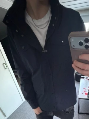 Svart vindjacka från bondelid - Säljer en svart vindjacka från Bondelid med klassisk krage och flera fickor framtill. Jackan har knappar och dragkedja samt en stilren design som passar perfekt till vardags. Materialet är polyester och jackan har långärmad passform.