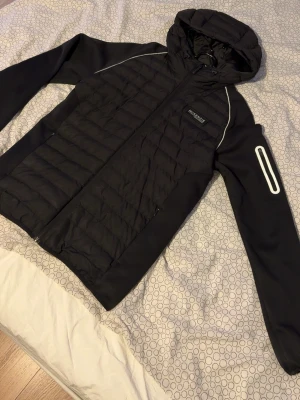 Svart vindjacka med huva McKenzie S - Svart cardigan från McKenzie med quiltad front, släta ärmar och reflekterande detaljer. Jackan har huva, dragkedja framtill och sportig look. Materialet är syntet och jackan har en modern passform som passar perfekt till streetwear.